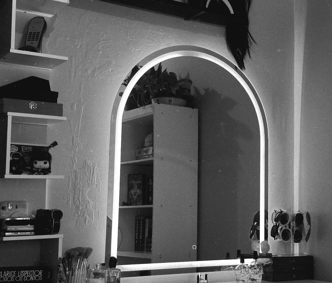 Local de atendimento da Maria em preto e branco: espelho em arco com luz de LED, prateleiras com livros e câmeras e pincéis de maquiagem na bancada